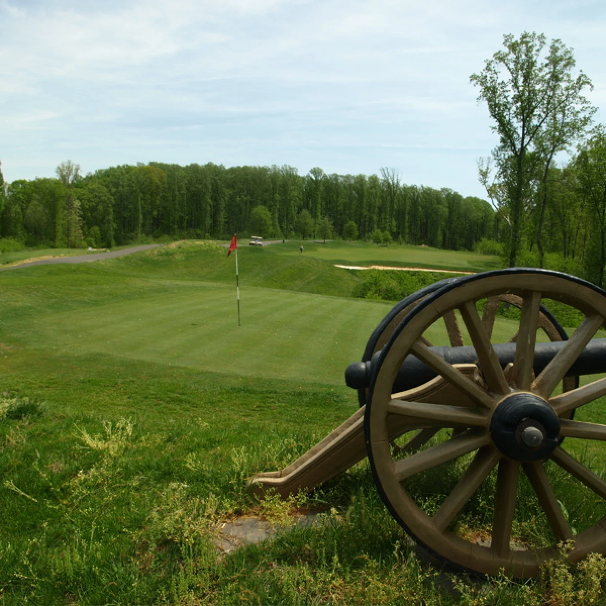 A cannon beside a golf green, with a red flag and wooded hills beyond.
