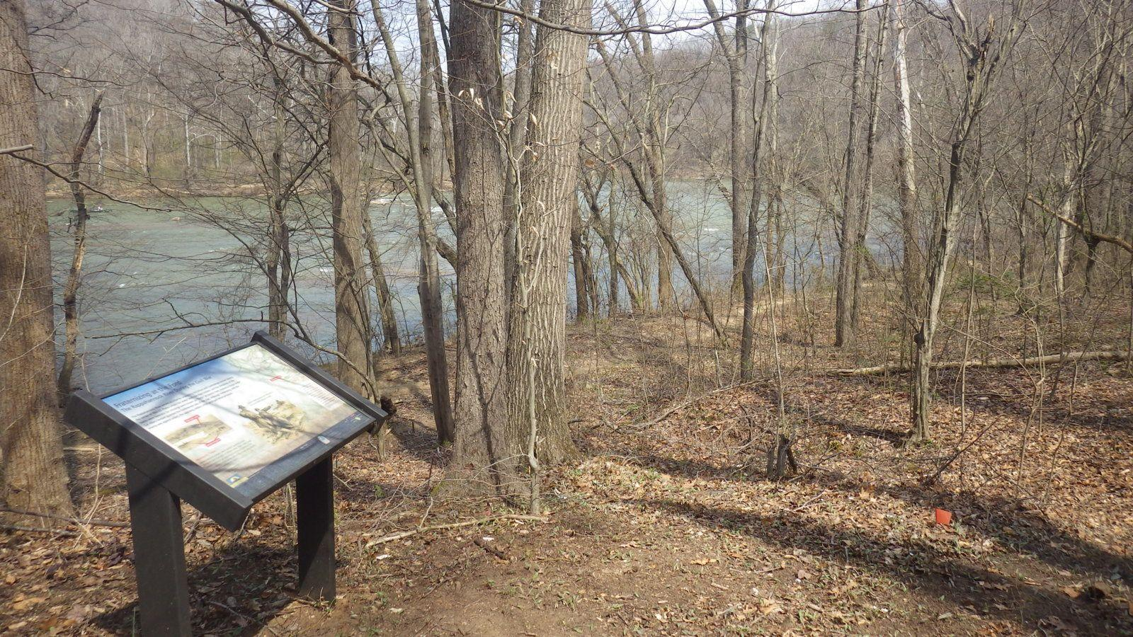 An interpretive panel stands among leafless trees beside a river.