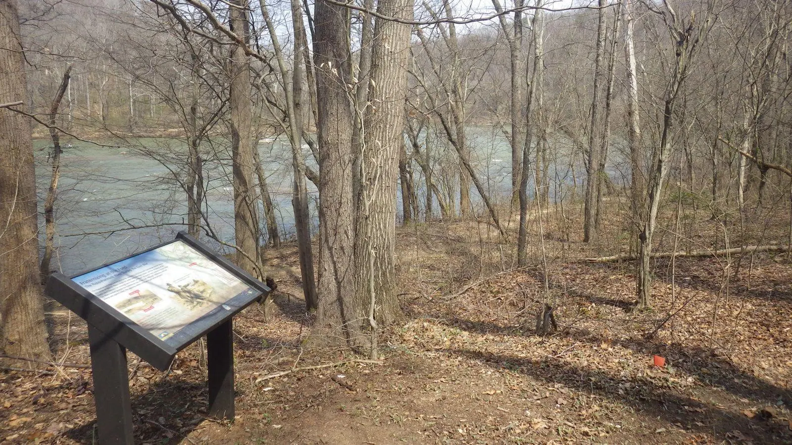 An interpretive panel stands among leafless trees beside a river.